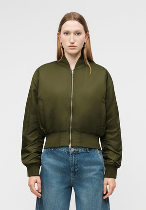 Γυναίκα με ίσια ανοιχτό καστανά μαλλιά, που φοράει κρίκους στα αυτιά, μια δερμάτινη μπορντό ζακέτα σε ελιά πράσινο χρώμα και μπλε τζιν ψηλόμεσο, μπροστά σε ένα απλό φόντο.