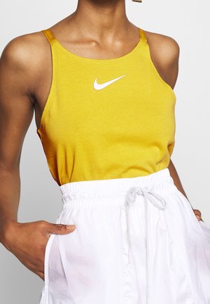 Una canotta gialla senza maniche presenta un logo Nike bianco, abbinata a pantaloni corte bianchi con cordino. Il tessuto appare morbido e di una consistenza liscia.
