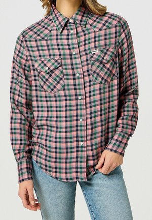 Kobieta ubrana w różową, zieloną i czarną koszulę w kratę zapinaną na guziki z dwoma przednimi kieszeniami oraz jasnoniebieskie dżinsy.