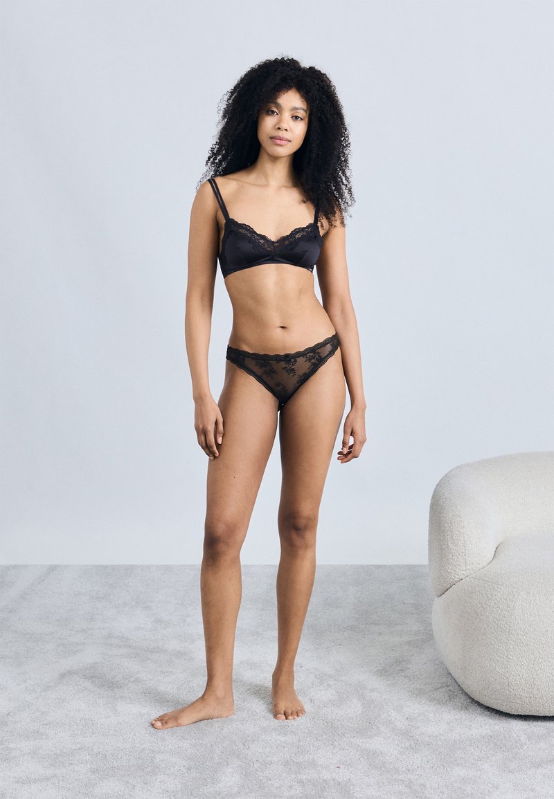 Musta pitsillä somistettu bralette ja string-sarja, jossa satiinimateriaali, säädettävät hihnat ja herkät kukkakuviot. Pehmeä, teksturoitu tausta.