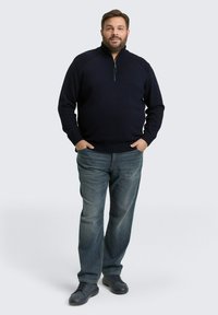 Marineblauwe pullover met een hoge kraag en ritssluiting, een gebreid ontwerp met textuur, gecombineerd met vervaagde blauwe jeans en donkere sneakers.