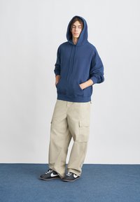 Navy blue cotton hoodie with a kangaroo pocket and drawstring, paired with beige cargo pants featuring side pockets. Worn with dark sneakers.