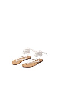 Sandali piatti bianchi con sottili cinturini, avvolti alla caviglia, e una suola di colore beige liscia. La suola presenta un design minimalista.