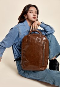 Smešani smeđi kožni ruksak s glatkom teksturom, prednji džep sa zatvaračem, dvostruke ručke i oznaka s logotipom. Postavljen na svijetlu pozadinu.