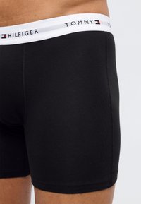 Κοντινό πλάνο ατόμου που φοράει μαύρα εσώρουχα μποξέρ με λευκή ελαστική μέση με την επιγραφή και το λογότυπο "Tommy Hilfiger".