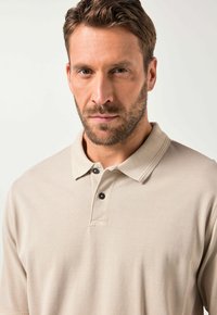 Beige pikétröja med strukturerad väv, två svarta knappar vid kragen och en något avslappnad passform. Korta ärmar och vikbar krage.