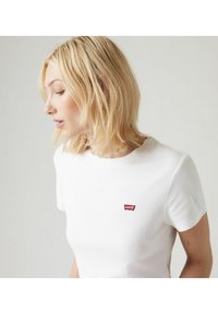 Camiseta blanca de algodón con mangas cortas, que presenta un pequeño parche de logo rojo de Levi's en el lado izquierdo del pecho. Diseño sencillo con una textura suave.