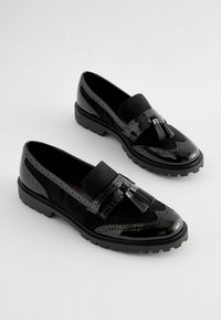Mocassini neri in una combinazione di pelle verniciata liscia e suede testurizzato. Accentuati da nappine e dettagli brogue, con suole robuste.