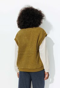 Gilet en polaire vert olive avec une surface douce et texturée, des manches courtes et un ourlet côtelé, porté par-dessus une chemise à rayures à manches longues.