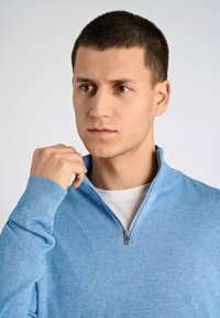 Jasnoniebieski sweter na zamek wykonany z miękkiej tkaniny, z prążkowanymi mankietami i zamkiem przy kołnierzu. Pod spodem biała koszulka.