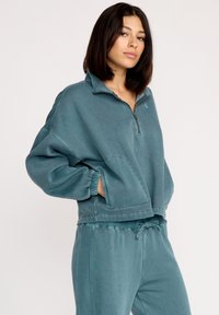 Teal cropped sweatshirt med halv-lynlås foran, lange ærmer og sidelommer. Har et blødt, struktureret stof og subtile syningsdetaljer.