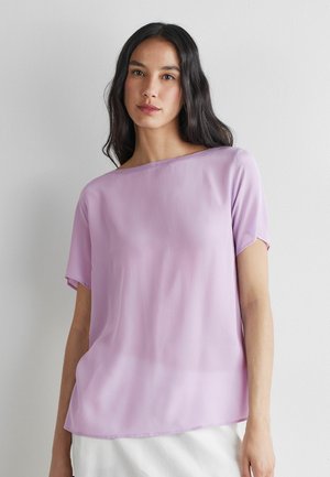 Vrouw met lang donker haar, die een losse, korte mouwen lavendelkleurige blouse en witte rok draagt, staand tegen een eenvoudige lichte muur.