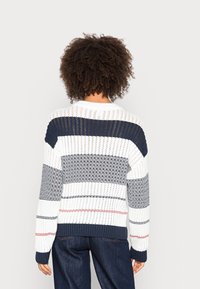 Personne aux cheveux bouclés portant un pull blanc en tricot avec des rayures horizontales bleu marine et rouges, vue de dos sur un fond uni.