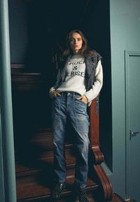 Maglione bianco con stampa nera "ROCK & DRISE", gilet grigio, jeans baggy blu, scarpe nere con borchie, sfondo con scala di legno, tonalità tenui.