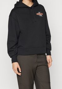 Persoon die een zwarte Calvin Klein Jeans-hoodie met trekkoorden en donkere bruine broek draagt, staand tegen een effen lichte achtergrond.