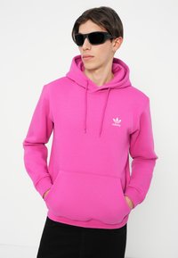 Ružová mikina s kapucňou a predným vreckom, s malým bielym logom Adidas. Model nosí čierne slnečné okuliare a tmavé nohavice.