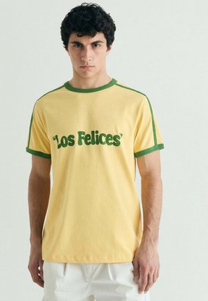 Joven con cabello corto, rizado y negro, que lleva una camiseta amarilla con ribetes verdes y el texto "Los Felices" en verde en el frente.