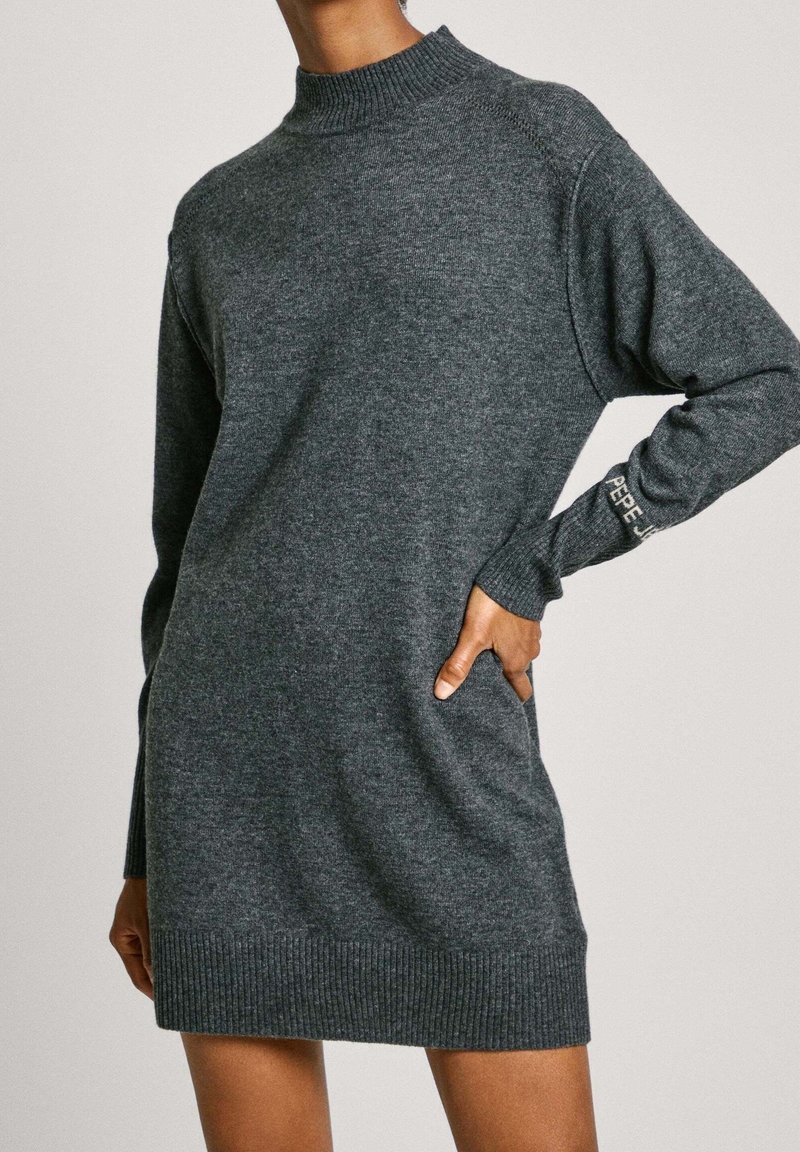 Robe-pull en maille gris foncé à manches longues avec col et ourlet côtelés, portée par une personne avec une main sur la hanche.