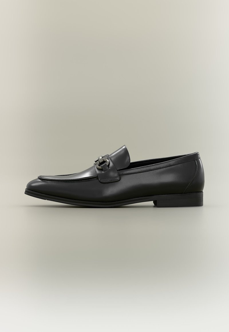 Mocassin pour homme en cuir noir avec un talon bas et un détail de boucle en métal sur le dessus, vu de profil sur un fond neutre.