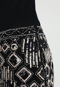 Schwarzer Stoff, verziert mit geometrischen Pailletten-Mustern in Schwarz, Silber und Creme, mit filigranen Designs und einem Reißverschluss.