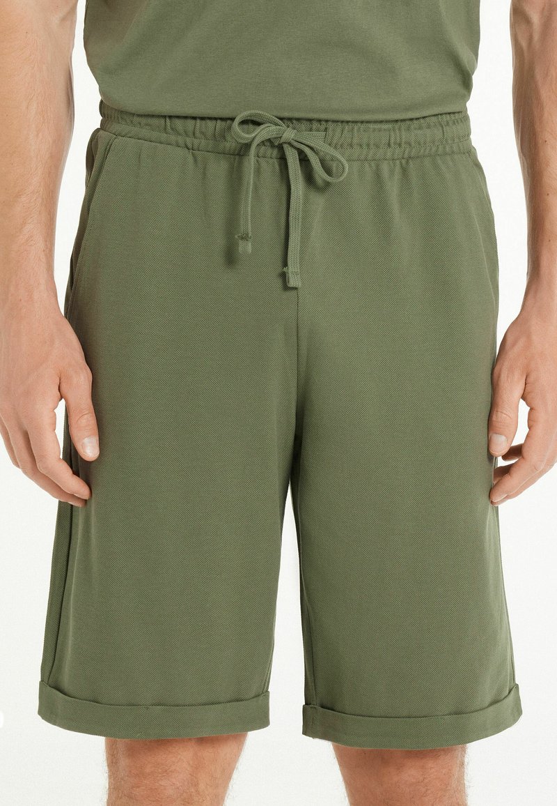 Pantalones cortos verde oliva hechos de una tela ligera, con una cintura elástica y cordón, y puños enrollados. Textura suave con un patrón sutil.