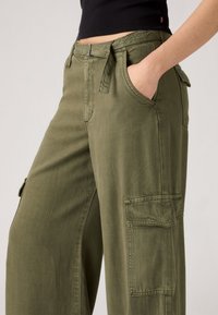 Persona che indossa pantaloni cargo verde oliva con tasche laterali e un crop top nero, con una mano in tasca, su uno sfondo semplice.