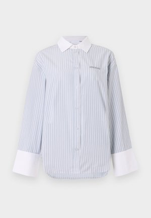 Camisa de botones a rayas en azul claro y blanco, con cuello y puños blancos. El material es suave, con un corte relajado y un sutil detalle de logo.