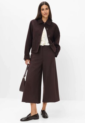 Kvinde iført mørkebrun cropped jakke, bukser med vide ben, hvid bluse med flæser, sorte loafers, holder en matchende brun håndtaske, står med hånden i lommen.