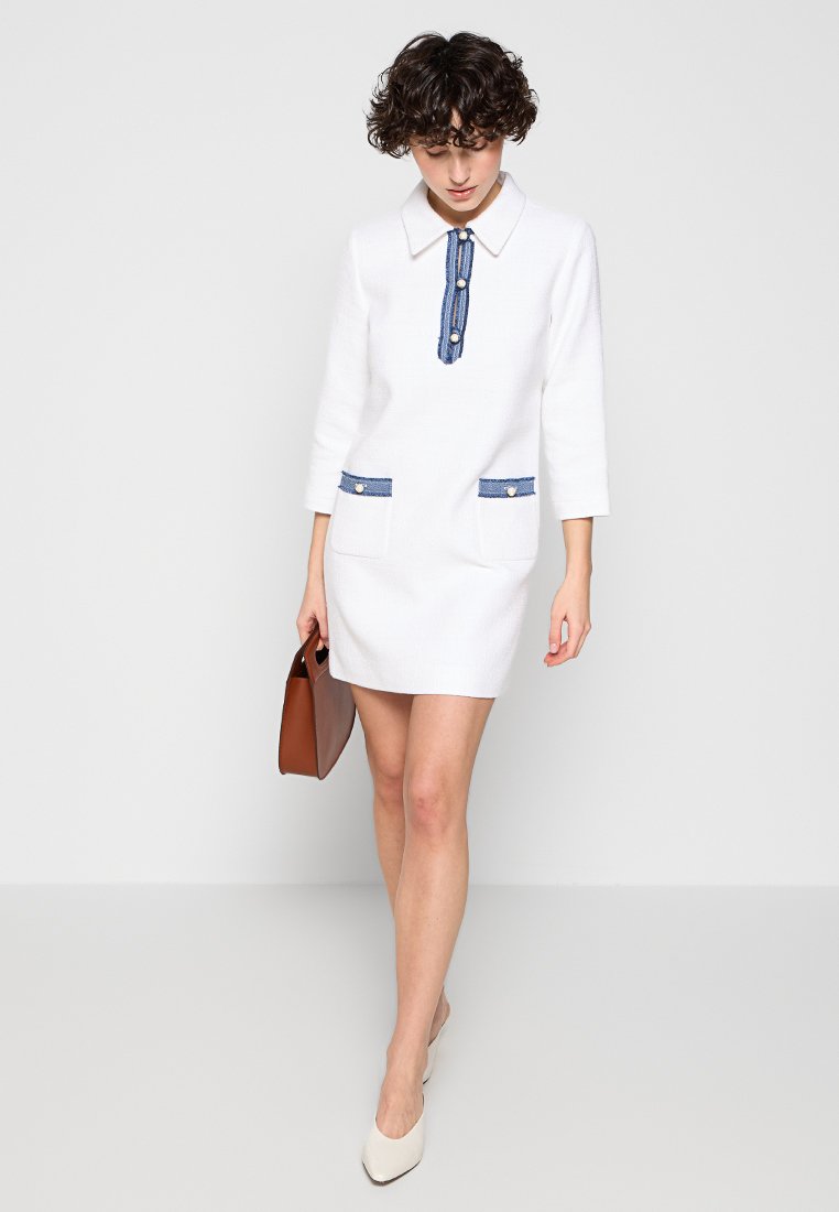 Weißes Kleid mit Dreiviertelärmeln, Denim-Akzenten an Taschen und Kragen, strukturierter Stoff und taillierter Schnitt. Model hält eine braune Tasche.