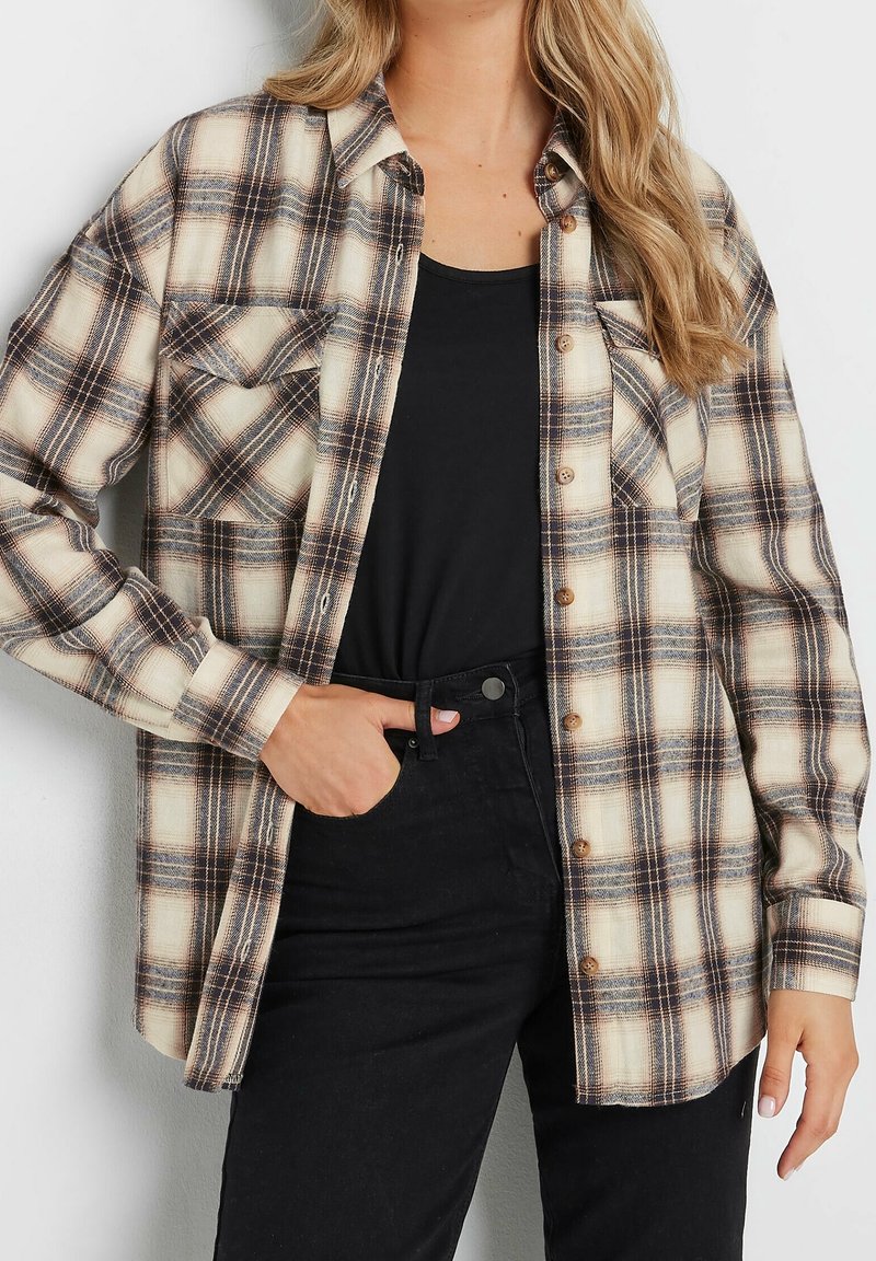Femme portant une chemise à carreaux beige et noire boutonnée sur un top noir et un pantalon noir, avec une main dans la poche.