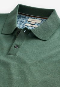 Tummanvihreä polo-paita, joka on valmistettu 100 % merinovillasta. Siinä on teksturoitu neule, kaulus ja kaksi mustaa nappia. Sisäetiketissä on geometrinen kuvio.