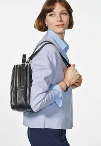 Czarny skórzany plecak z srebrnymi zamkami, miękka tekstura, zaokrąglony kształt, noszony na jasnoniebieskim kardiganie i koszuli.