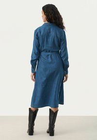 Denim haljina, duge rukave, zatvoreni gumbaste prednje strane, s remenom u struku, dužine do sredine lista, duboke plave boje, lagane teksture, uparena s crnim čizmama do gležnja.