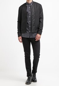 Giubbotto bomber nero con una texture liscia, chiusura con zip e colletto rotondo, abbinato a una camicia button-up grigio scuro, pantaloni slim-fit neri e stivali.