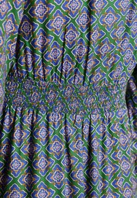 Bavlnená tkanina so vzorom má zelené pozadie s modrými, bielymi a zlatými geometrickými motívmi. Zhromaždený pás s detailom smock dodáva textúru.