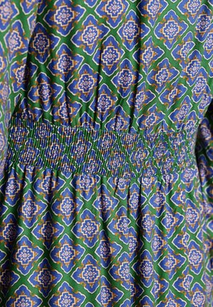 Měkoučká bavlněná tkanina s vzorem má zelené pozadí a modré, bílé a zlaté geometrické motivy. Ujímaný detail na pasu dodává texturu.