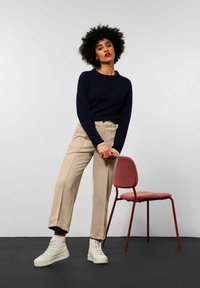 Maglione blu navy, pantaloni beige su misura e sneakers alte bianche. Sedia di metallo rossa sullo sfondo con seduta rosa. Design minimalista e palette di colori.