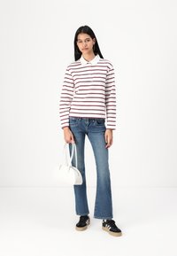 White long-sleeve polo with red stripes, blue flared jeans, black sneakers, and a white handbag. Model stands against a plain background.