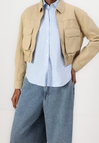 Henkilö, jolla on beige lyhyt takki sinivalkoraidallisen napitettavan paidan päällä ja löysät vaaleansiniset farkut, käsi taskussa.