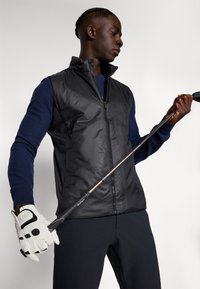 Čierna polstrovaná vesta s vysokým golierom, nosená cez námornícku dlhú košeľu. Model drží golfovú palicu, oblečenú v bielej rukavici s čiernymi akcentmi.