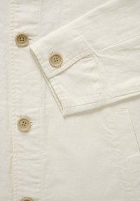 Gros plan sur une veste en tissu crème montrant le devant boutonné et le poignet avec des boutons ronds beige.