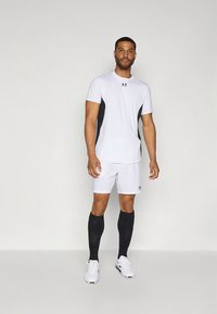 Camisola desportiva branca com painéis laterais pretos, combinada com calções brancos e meias pretas até ao joelho. Apresenta o logótipo da Under Armour na camisola e nos calções.