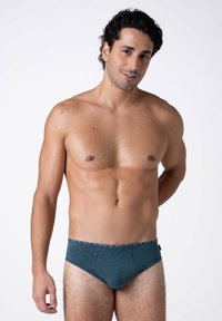 Slip in cotone verde con una cintura a righe orizzontali, design aderente, texture liscia e taglio basso, indossato da un uomo.