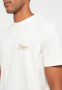 Camiseta de algodón blanca con una textura suave, que presenta un logo bordado en beige en el lado izquierdo del pecho y un cuello redondo clásico.