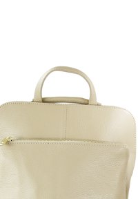Sac à dos en cuir texturé beige avec une poignée supérieure, une poche frontale zippée et des détails de couture visibles. Forme compacte et structurée.