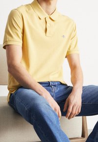 Gul polo-shirt med krage och två knappar, tillverkad av bomullsblandning. Bärs med blå denimjeans, sittande på en stol i neutral färg.