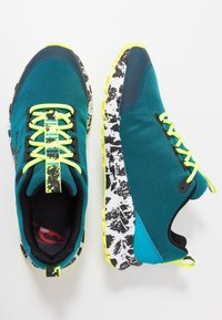 Chaussures de sport en maille bleu sarcelle avec semelles à motifs noirs et blancs, accents jaunes et lacets noirs. Bout arrondi et col rembourré.