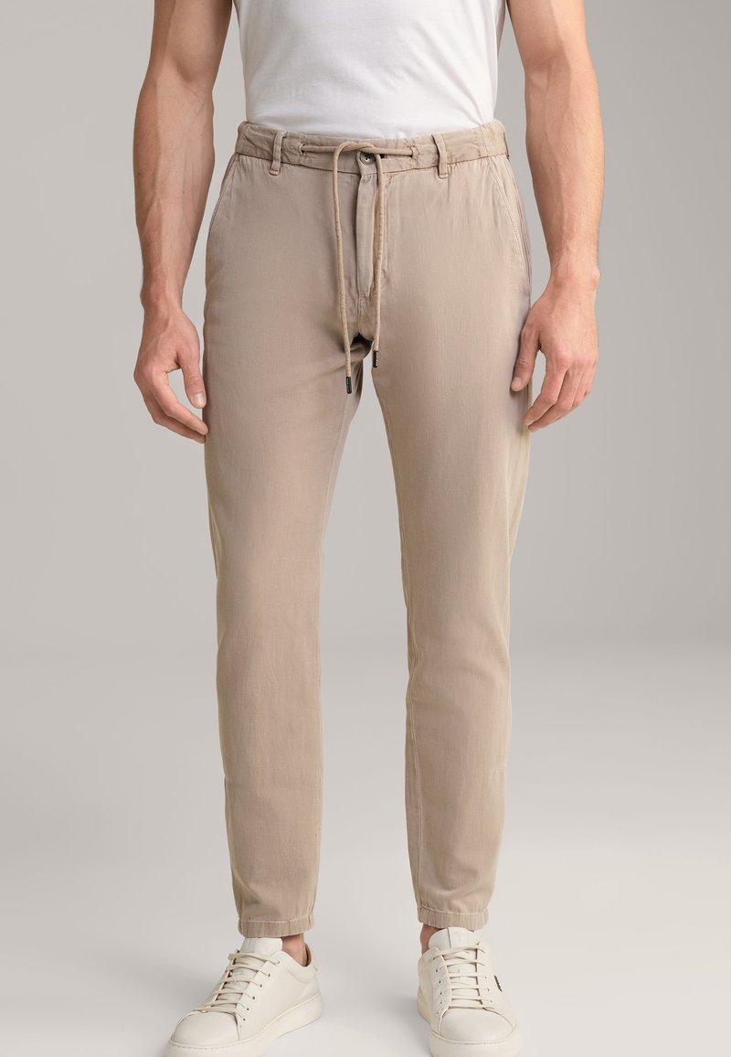 Homme portant un pantalon décontracté beige avec cordon de serrage et des baskets blanches, debout devant un fond uni.