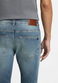 Jeans de mezclilla azul claro con acabado desgastado, que cuentan con una etiqueta de cuero marrón que dice "BOSS", diseño de cinco bolsillos y detalles de costura.