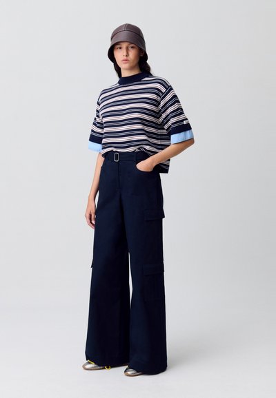 Camisa de manga corta de rayas en azul marino, crema y azul claro; pantalones cargo anchos en azul marino con bolsillos; gorra de cubo gris; zapatillas plateadas con acentos amarillos.